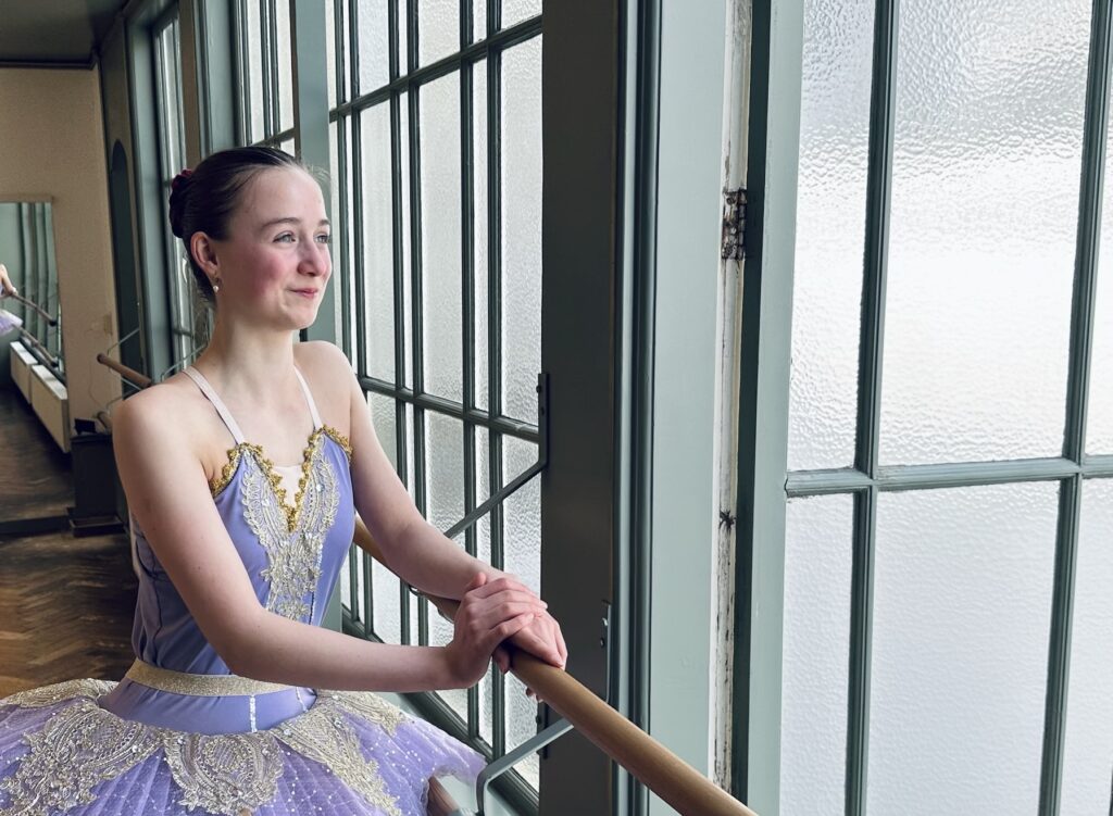 Balletstudent in tutu leunt dromerig op de barre met een glimlach en kijkt uit het raam, een fijn vooruitzicht.